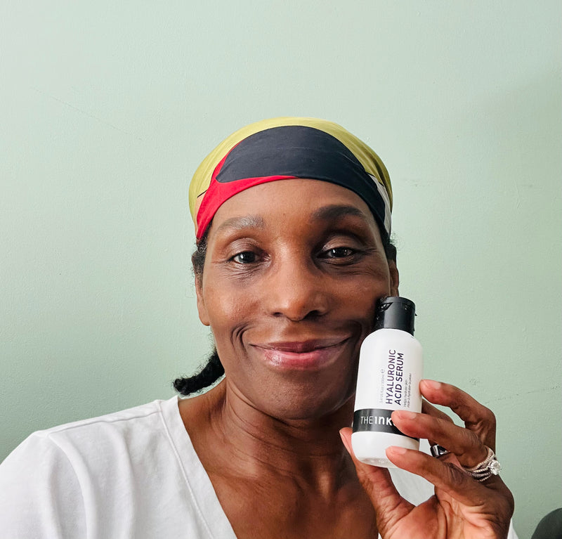 A woman wearing a black, yellow, and red headband holds up a white bottle of The Inkey List 'Hyaluronic Acid Serum' against a light mint green wall.