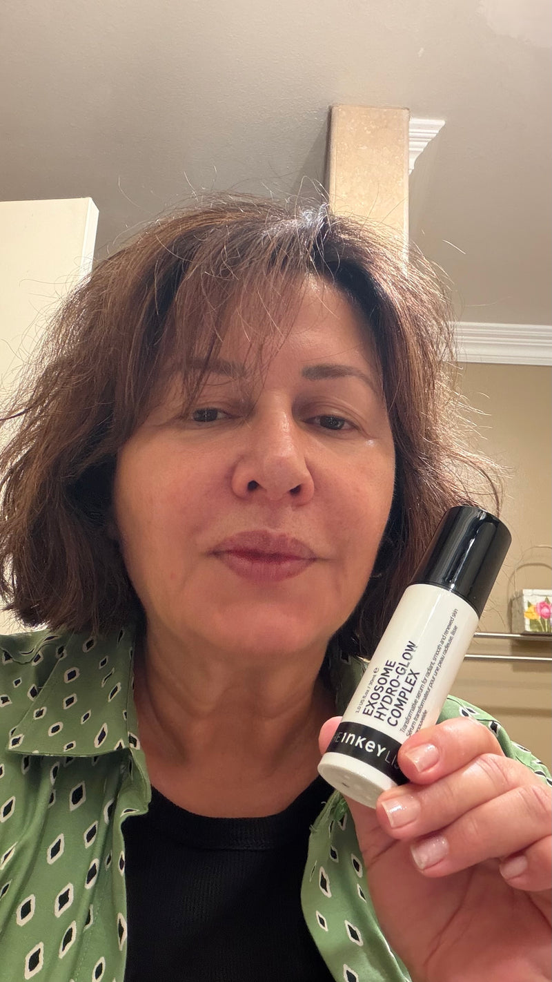 A woman holding a white and black bottle of Exosome Serum in front of her face, set against an indoor home background.