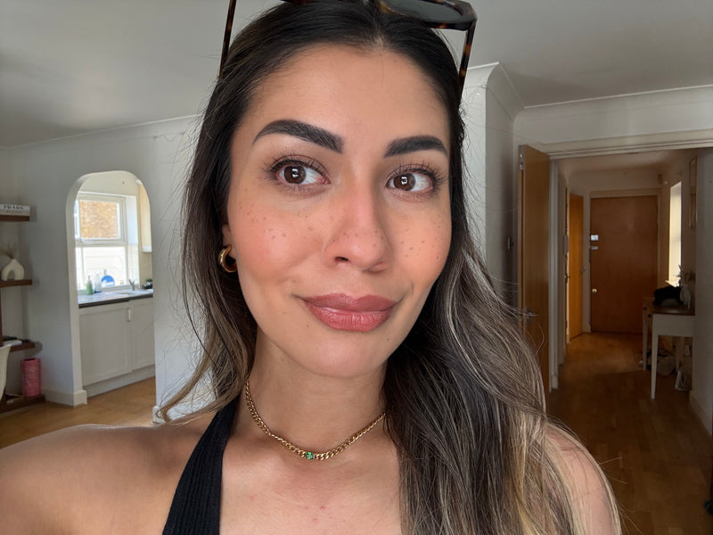 A close-up selfie of a woman with dramatic black winged eyeliner and dark eyebrows, wearing a black top and gold chain necklace in an indoor setting.