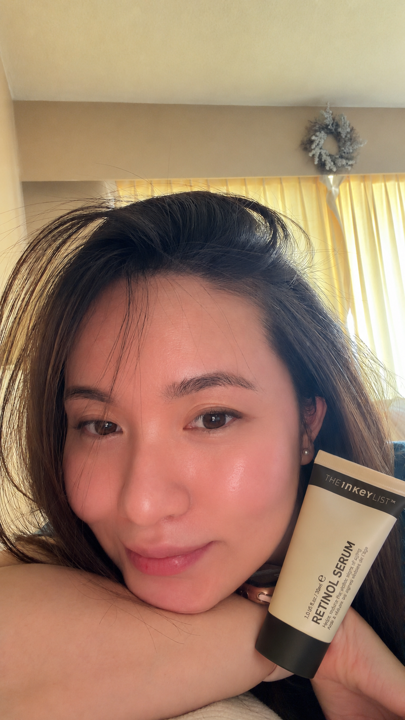 A woman holding a tube of INKEY list Retinol Serum in front of her face in an indoor setting with warm yellow curtains in the background.
