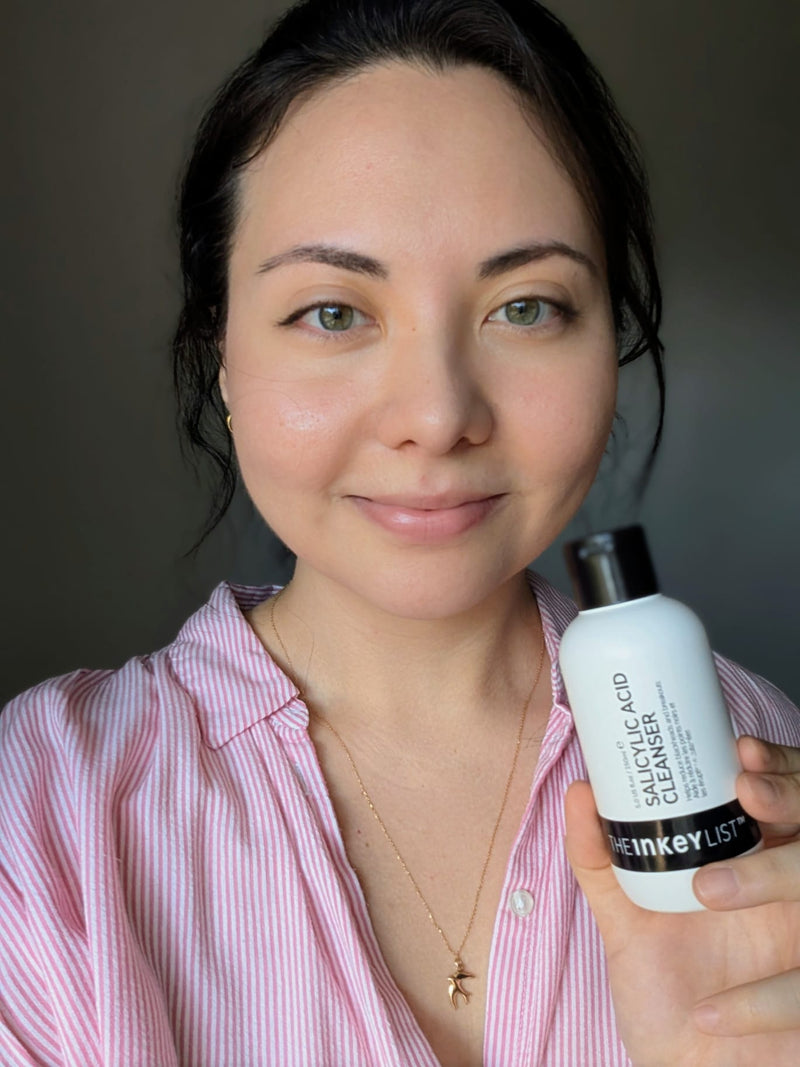 A woman with dark hair pulled back, wearing a pink and white striped button-up shirt, holding a white bottle of 'The Inkey List' Salicylic Acid Cleansing Foam against a dark gray background.