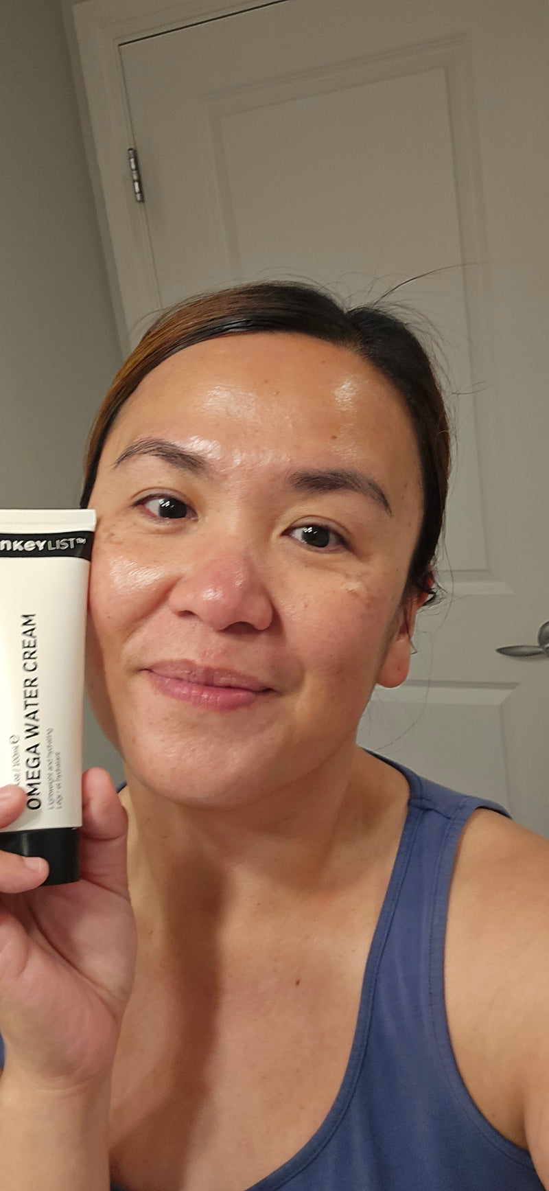 A woman holding a tube of Omega Water Cream near her face in a bathroom setting.