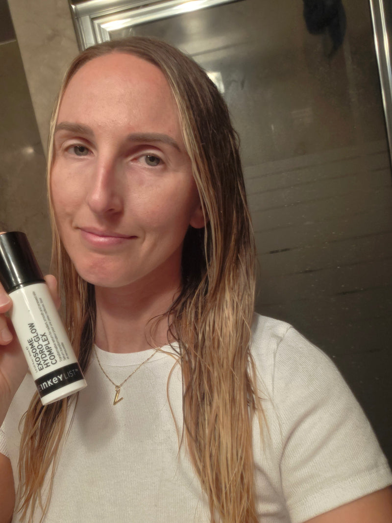 A woman holding a white bottle of Exosome Glow Complex in front of her face, with her long blonde hair visible in a bathroom setting.