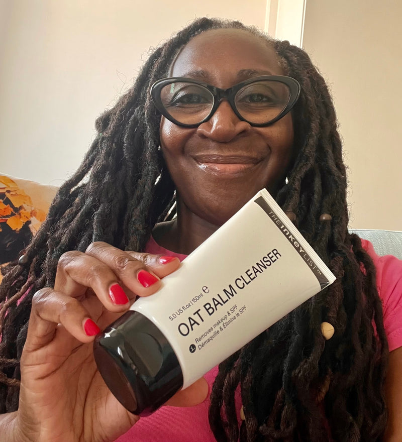 A smiling woman with long black hair and glasses holds up a white tube of Oat Balm Cleanser. The product packaging is white with black text and a black cap, and the woman has red nail polish.