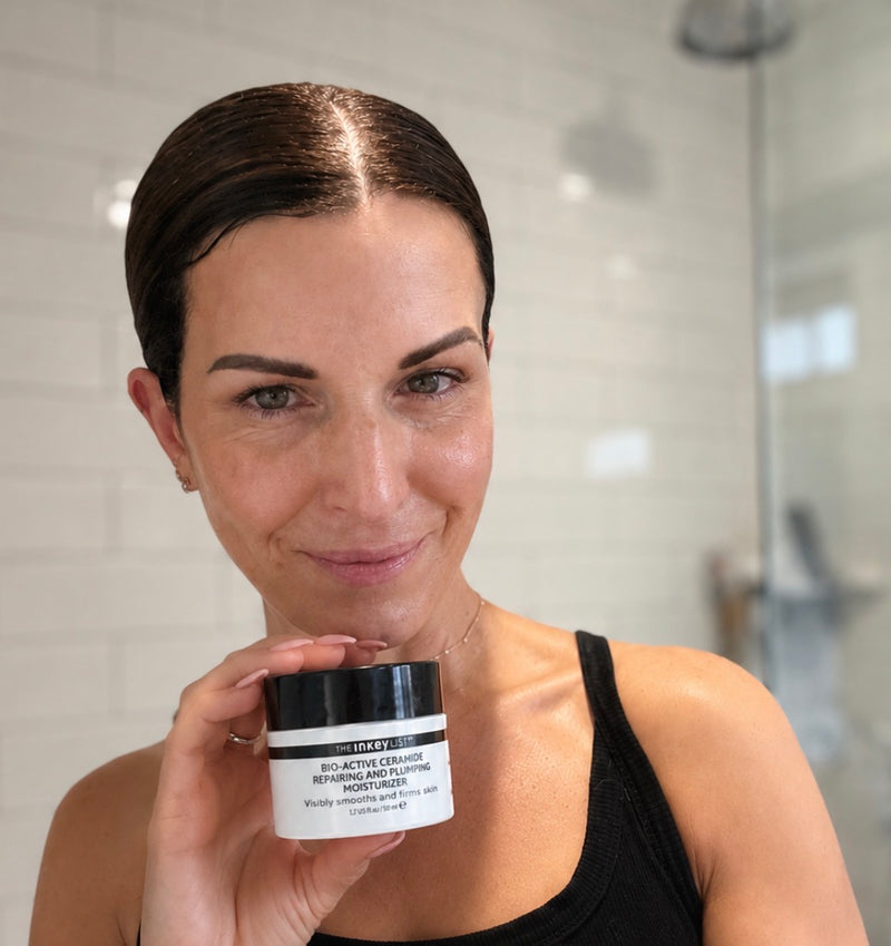 A woman in a black tank top holds a jar of Bio Active moisturiser in a bright bathroom setting with white subway tiles.
