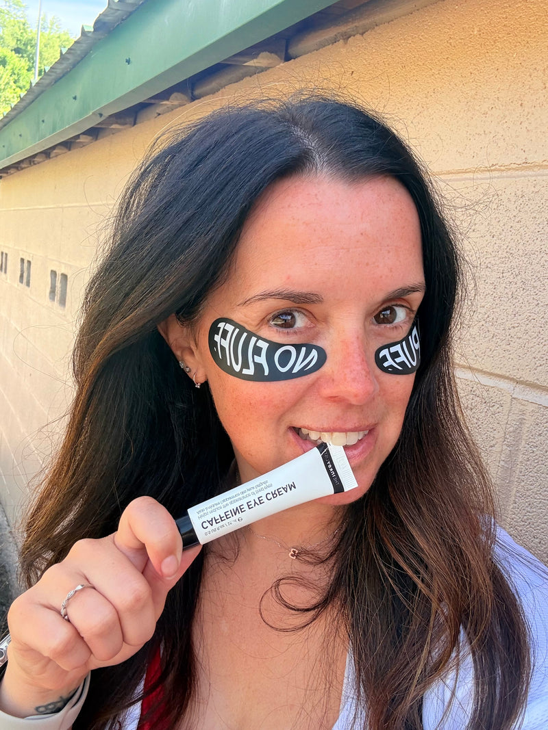 A woman holding a tube of Caffeine Eye cream in front of her face, and she has black eye patches with white text. She is standing outdoors against a beige stone wall.