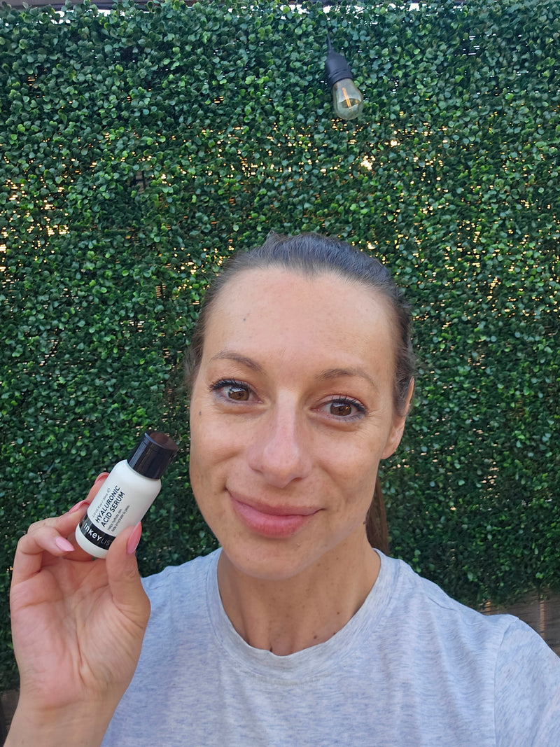 A woman holding a small white cosmetic bottle with a black cap, photographed outdoors against a dense green hedge background.