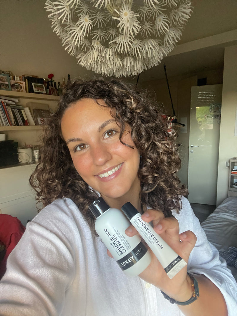 A smiling woman with curly brown hair holds up two skincare products, a white bottle labeled 'Glow Serum' and a white tube labeled 'Eye Cream', in a selfie-style photo taken in a bedroom with a decorative hanging light overhead.