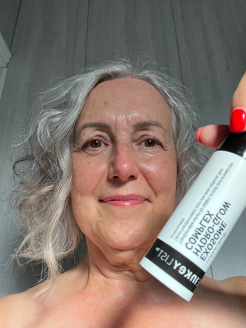 A close-up selfie of a woman with gray hair holding a white bottle of Exosome against a gray paneled background.