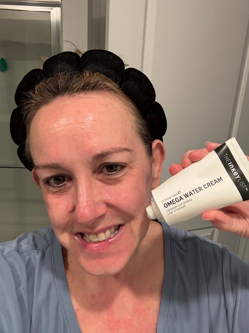 A woman wearing a black scrunchie headband and blue top holds up a white tube of 'The Inkey list' Omega Water Cream in a bathroom setting.