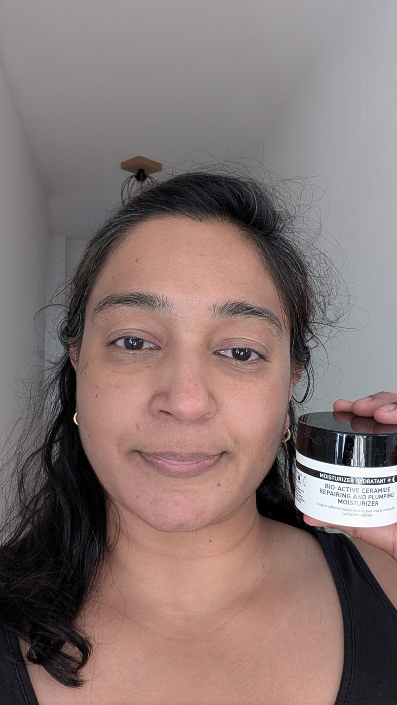 A woman holding a jar of face cream in a home setting with a white wall and ceiling visible.