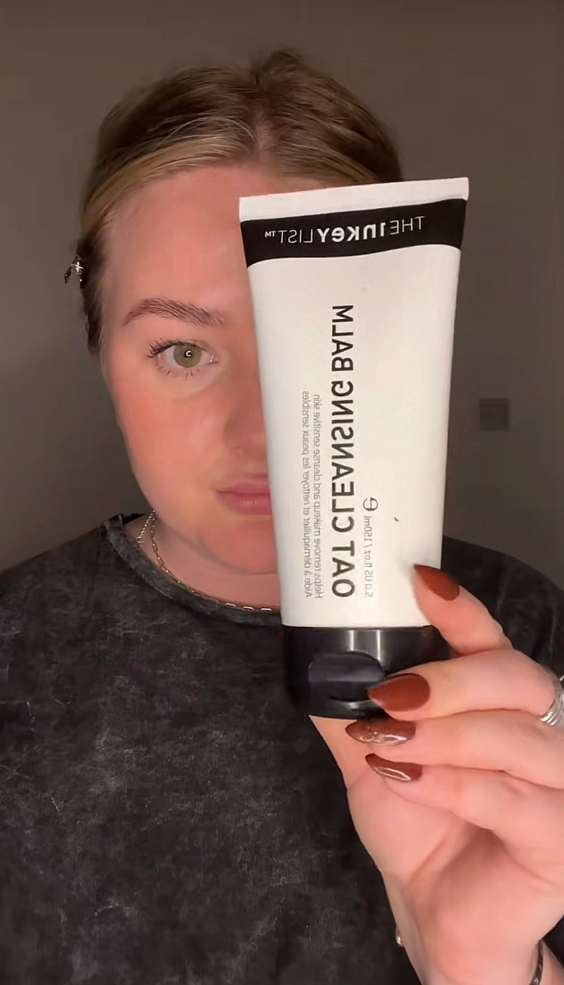 A woman holds up a white tube of Oat Cleansing Balm in front of her face, showing the product in a close-up shot against a plain gray wall.