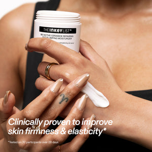 A close-up of a person applying white cream from The Inkey List Bioactive Cleansing Repairing Hand in Tanning Moisturizer onto their hand. The person is wearing beige manicured nails and gold rings, and the background is a soft-focus view of their bare torso.