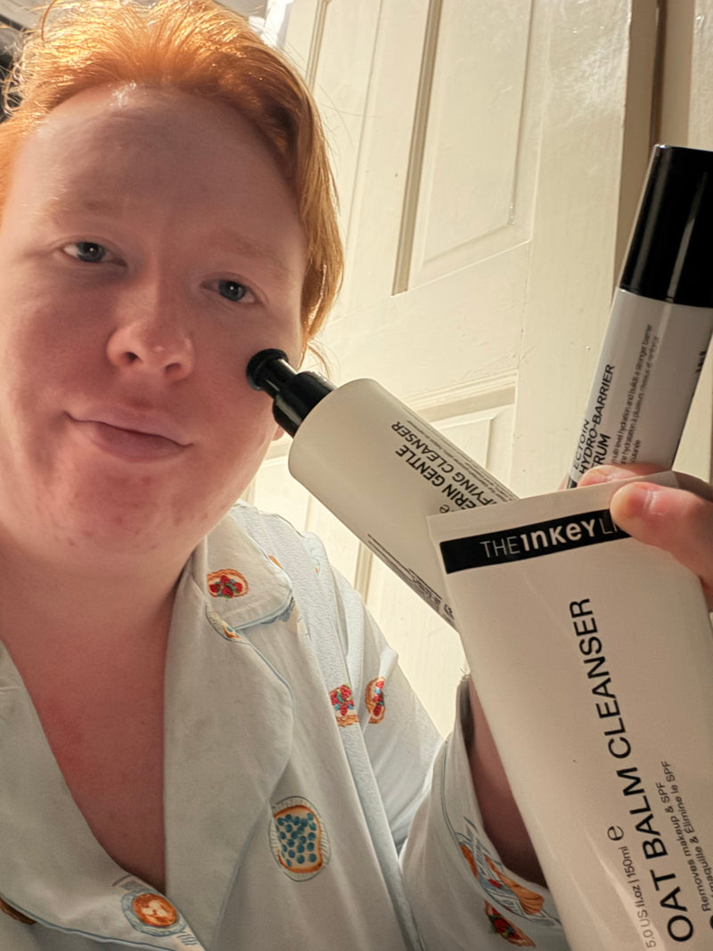 A person in a light blue pajama shirt holds up skincare products, including a white tube labeled 'THE INKEY OAT BALM CLEANSER' in front of a white paneled door.