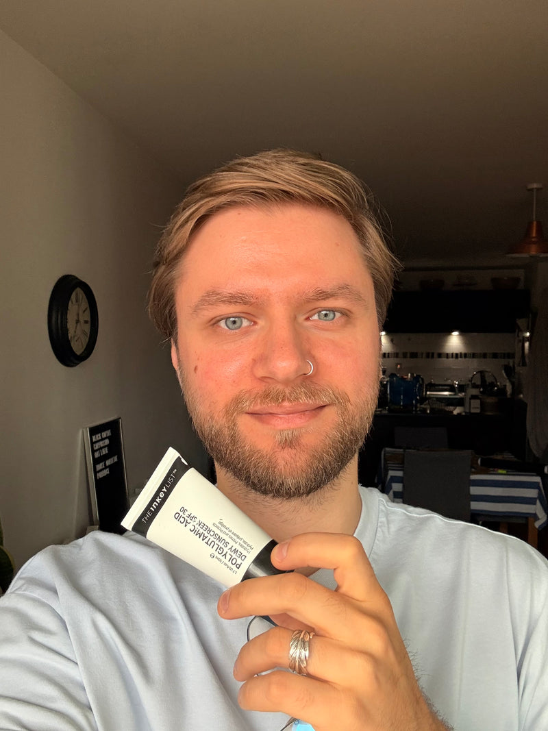 A man holding a white and black tube of SPF face wash in a home setting.