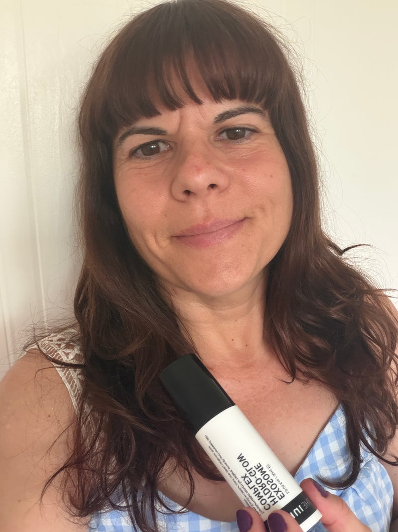 A woman with brown hair holding a white and black exosome serum bottle against a plain off-white wall background.