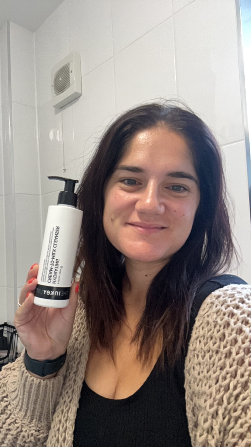 A woman holding a white pump bottle of The Inkey List Hydrating Cream-to-Milk Cleanser in a bathroom with white tiled walls.