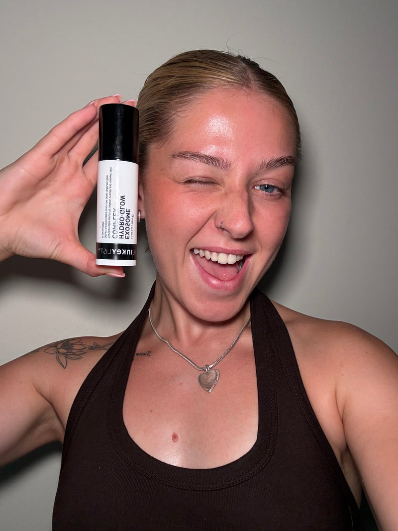 A woman holding up a white and black bottle of Exosome Serum against a plain gray wall.