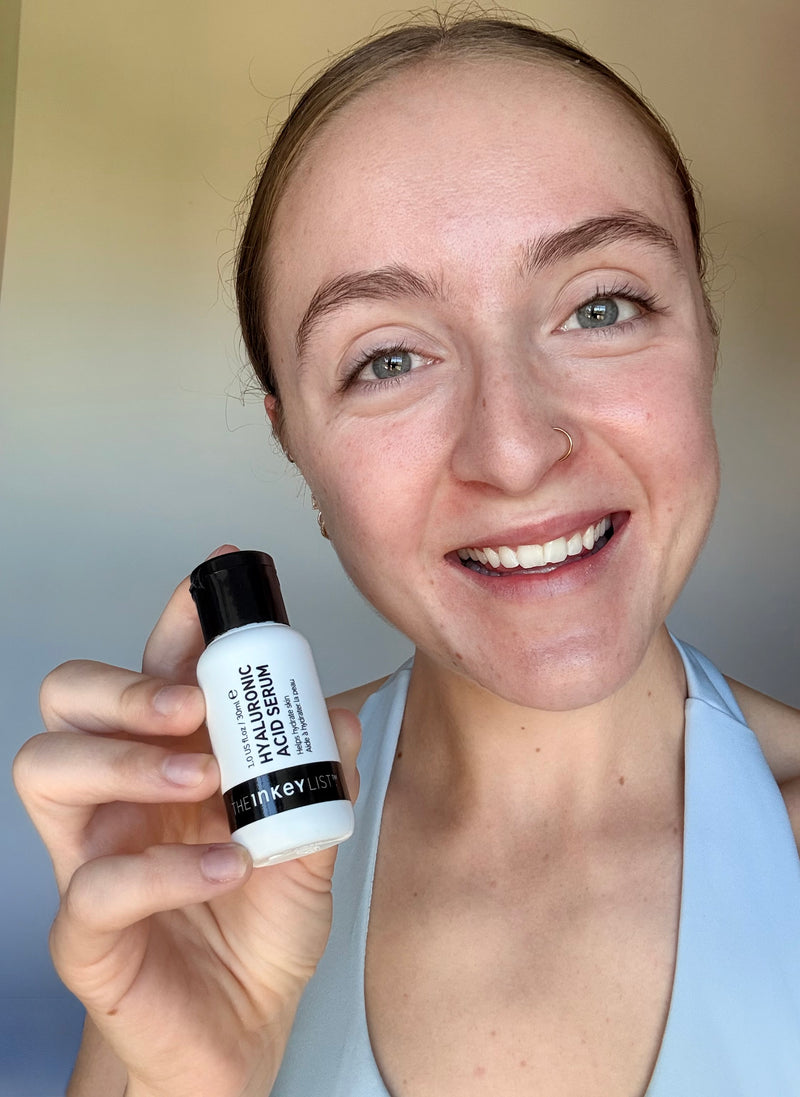 A smiling woman holds up a small white bottle of The Inkey List Hyaluronic Acid against a soft beige background.