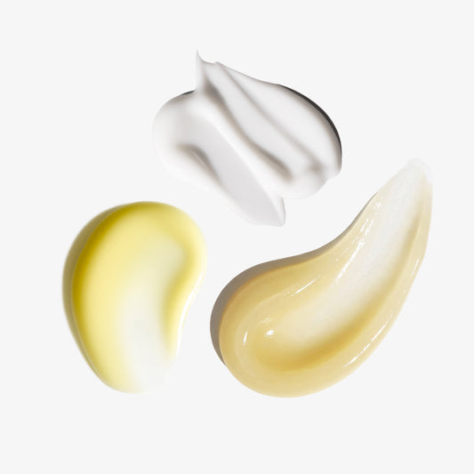 Three cosmetic cream swatches on a light gray background: a thick white textured swipe at the top, a smooth pale yellow oval at lower left, and a glossy translucent beige teardrop at lower right.