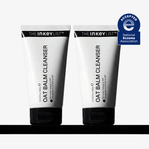 Two white squeeze tubes of THE INKEY LIST OAT Balm Cleanser with black caps, displayed side-by-side on a black surface against a light gray background, with a blue National Eczema Association Accepted badge in the top right corner.