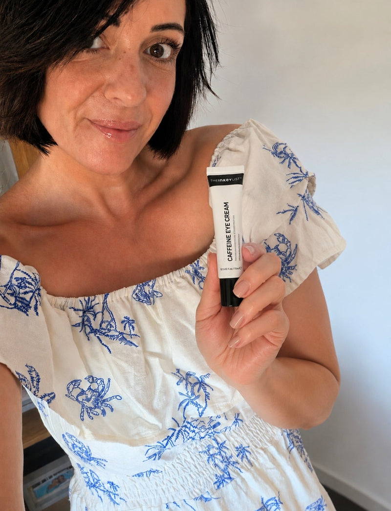 A woman holding a black and white tube of 'Caffeine eye cream', wearing a white off-the-shoulder dress with a blue floral pattern.