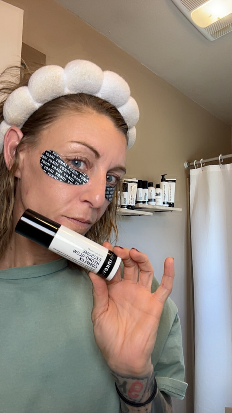 A woman in a bathroom wearing a white pom-pom headband and eye patches, holding a black and white cosmetic tube labeled 'Exosome' near her face.