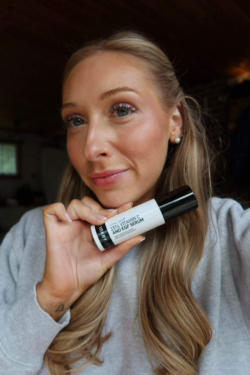 A woman with blonde hair holding a white and black tube of 15% Vit C, wearing a light gray sweatshirt in an indoor setting.