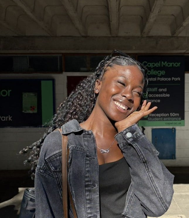 A smiling woman with long black curly hair posing outdoors near a station or entrance, wearing a dark denim jacket over a black top and carrying a shoulder bag.
