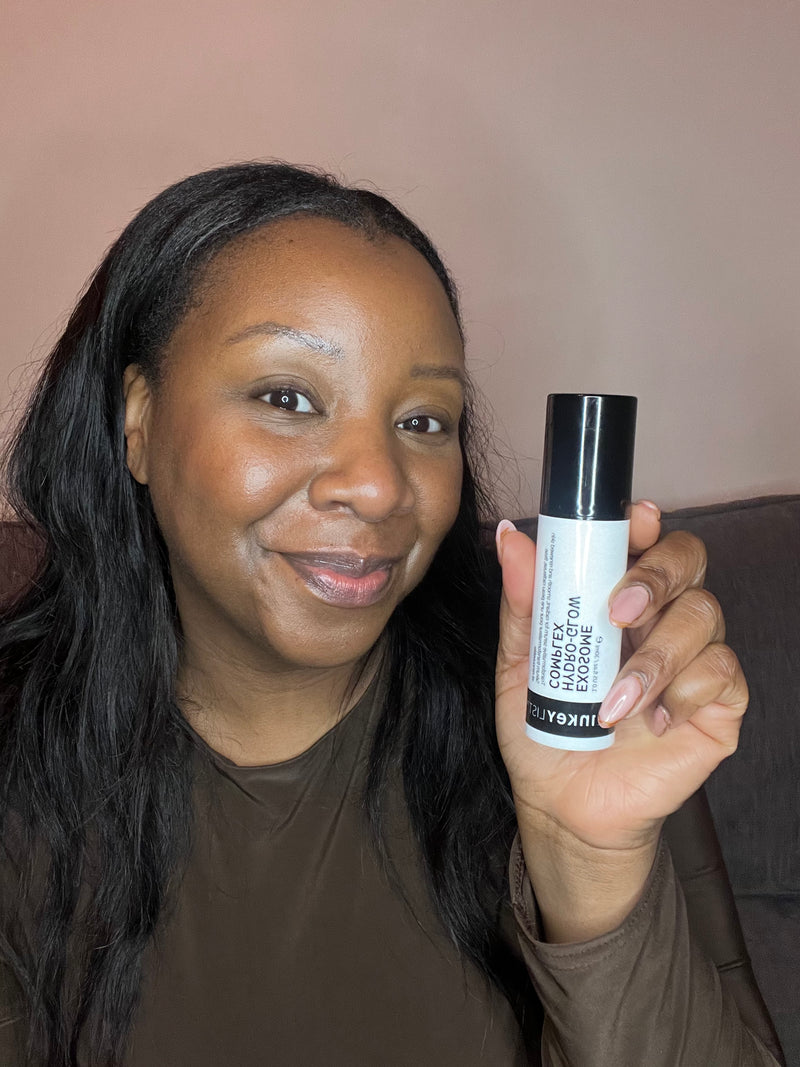 A woman holding a black and white Exosome serum in front of her face, wearing a brown long-sleeve top against a plain beige background.