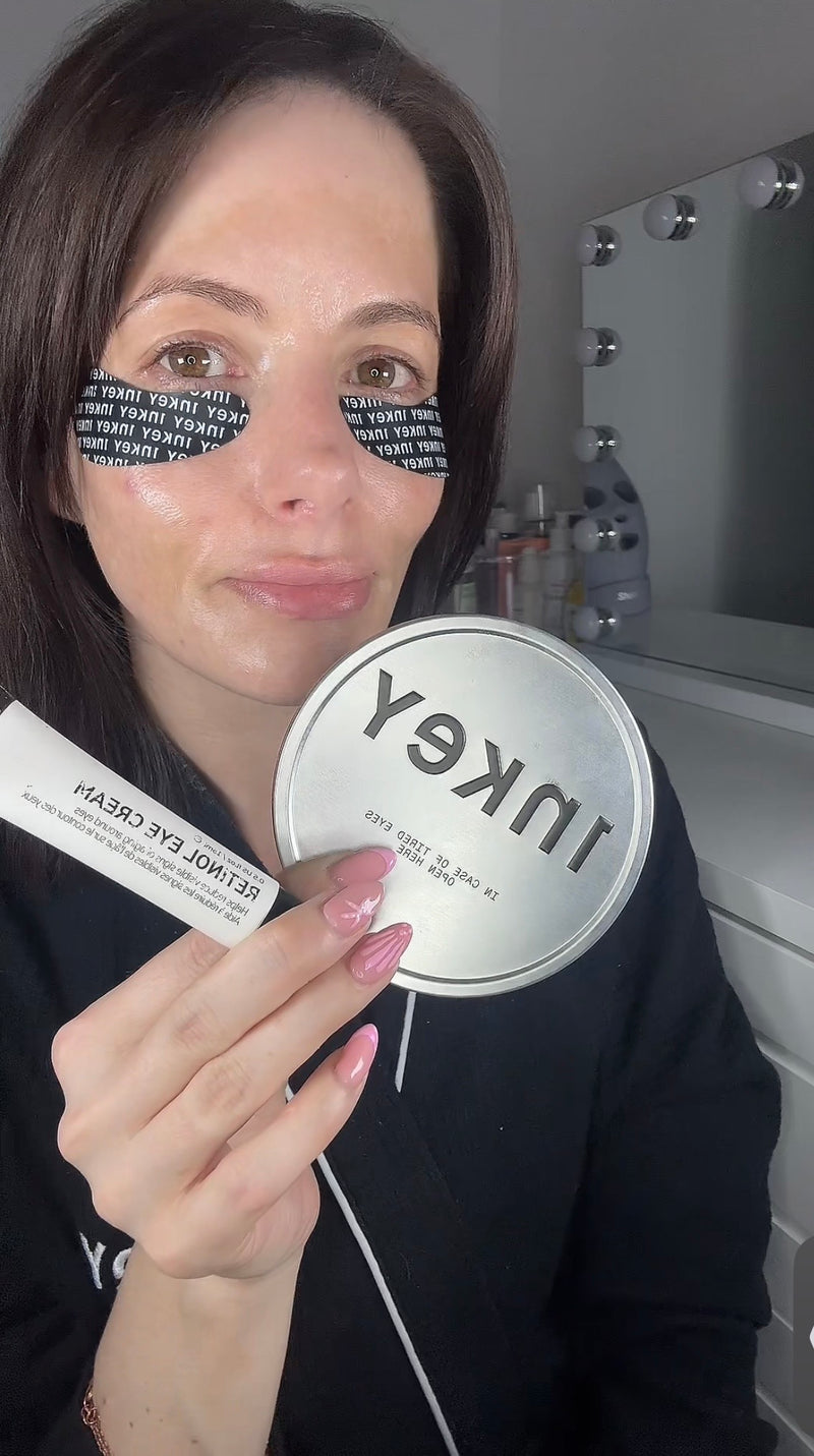 A woman holding a round silver tin and a white tube of Inkey Retinol Eye Cream in front of her face, with black eye patches on her eyes and a vanity mirror in the background.