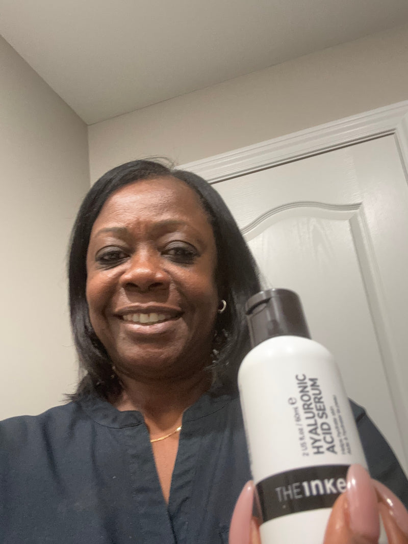 A woman holding a white bottle of The Inkey List Serum in front of a white door and beige wall.
